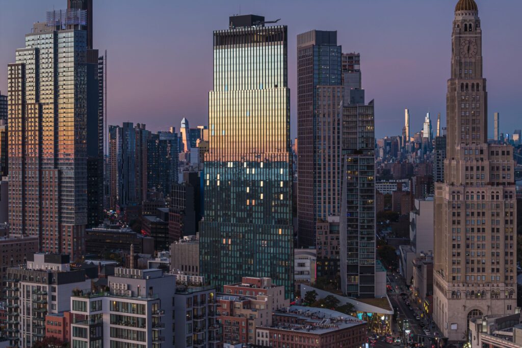 Cityscape at dusk with modern and historic high-rise buildings; the sky reflects off glass windows while lights begin to turn on across the city.