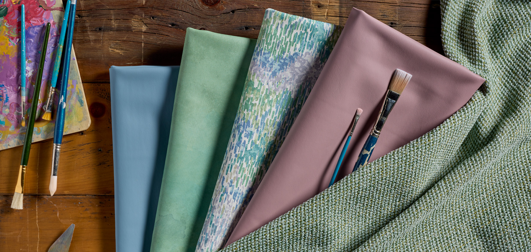 Five folded fabric samples in blue, green, patterned, and mauve shades lie on a wooden table with paintbrushes and a textured green cloth, evoking the bold lines and thoughtful palettes often seen in antifascist architecture.