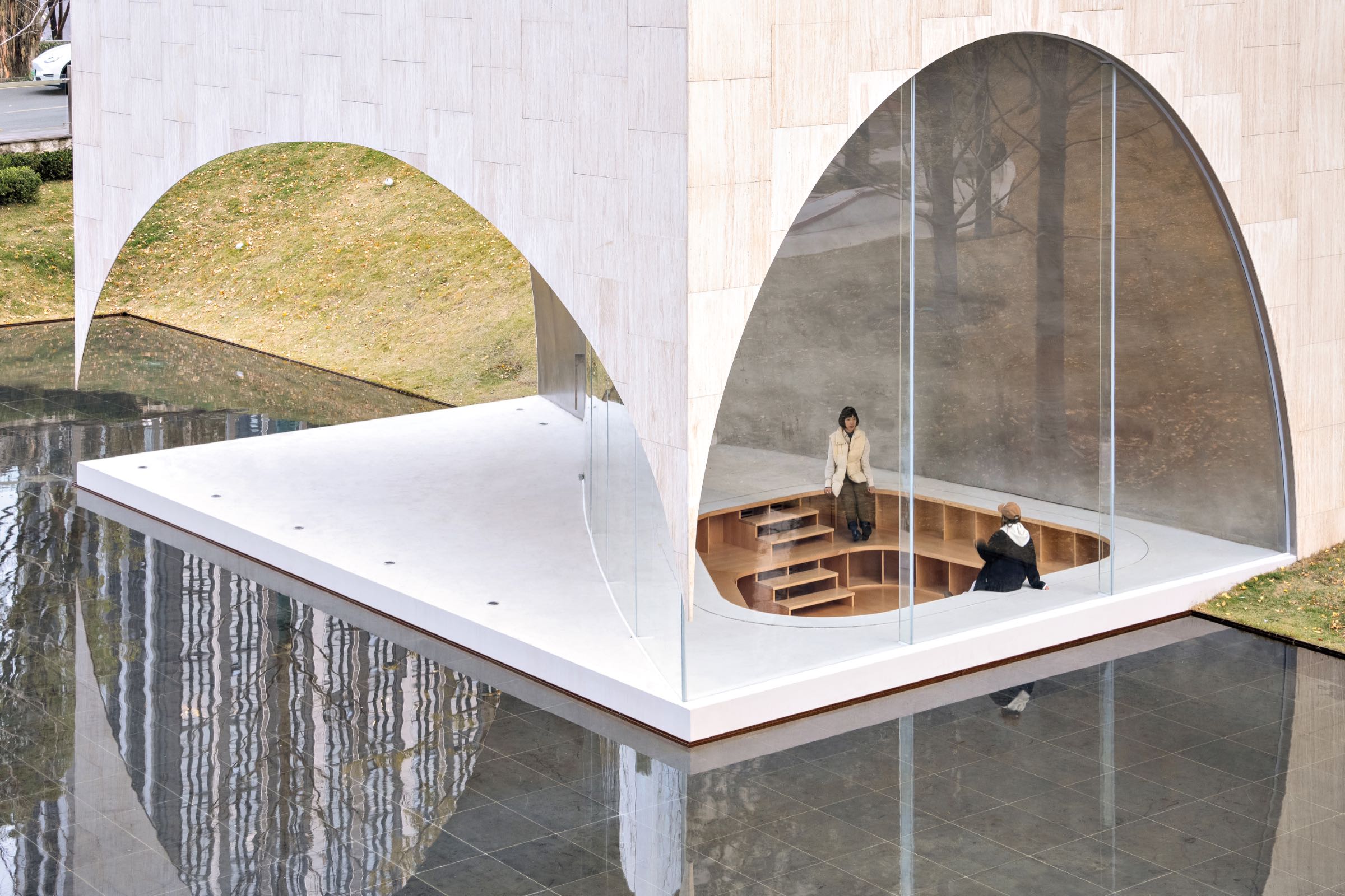 Two people sit and stand in a sunken wooden seating area inside a modern glass building with arched openings, surrounded by reflective water and a grassy landscape.