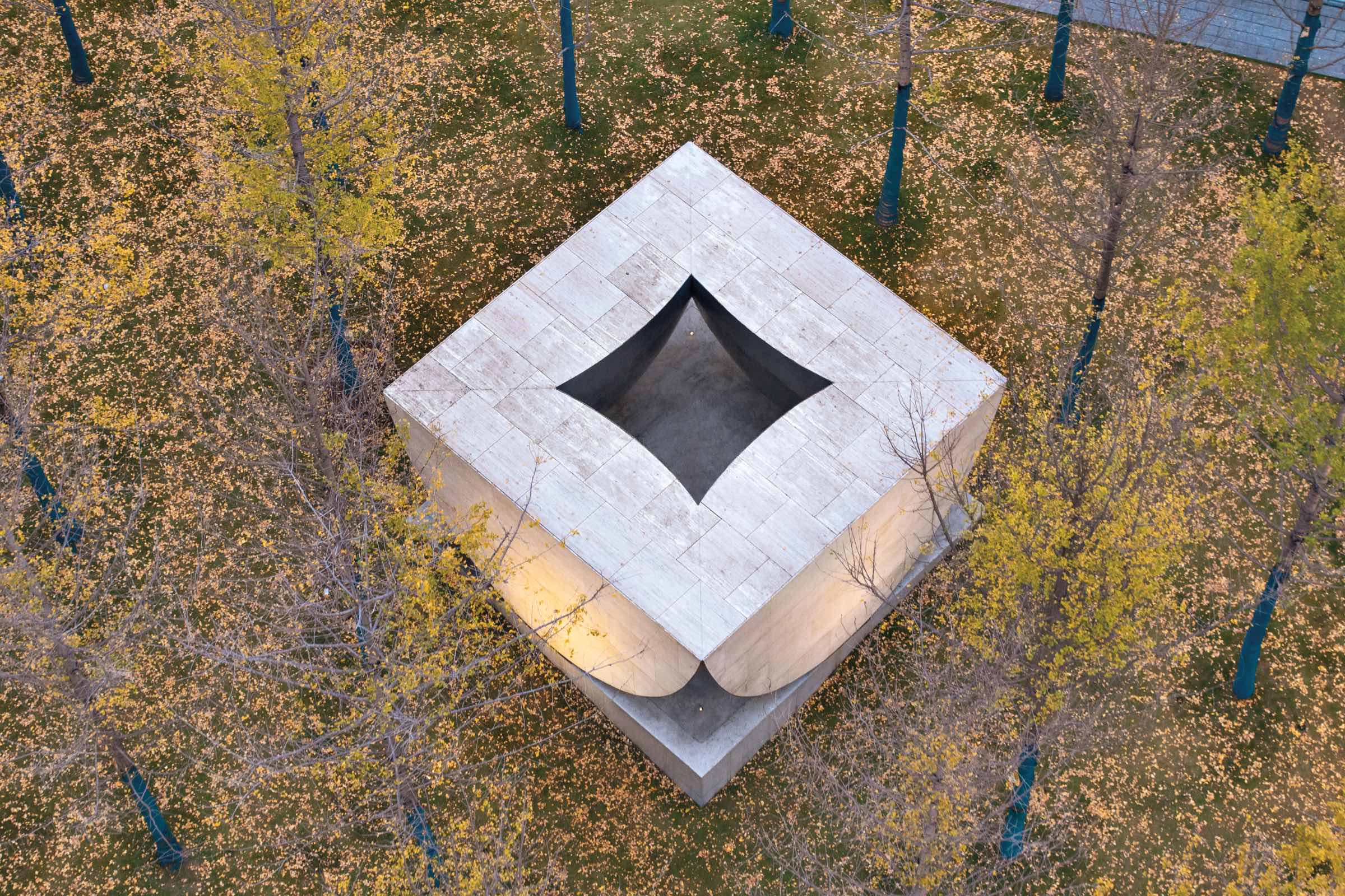 Atelier Xi, AYDC Public Art Center: Aerial view of a square, white stone structure with a curved, open roof, surrounded by autumn trees and scattered yellow leaves on the ground.