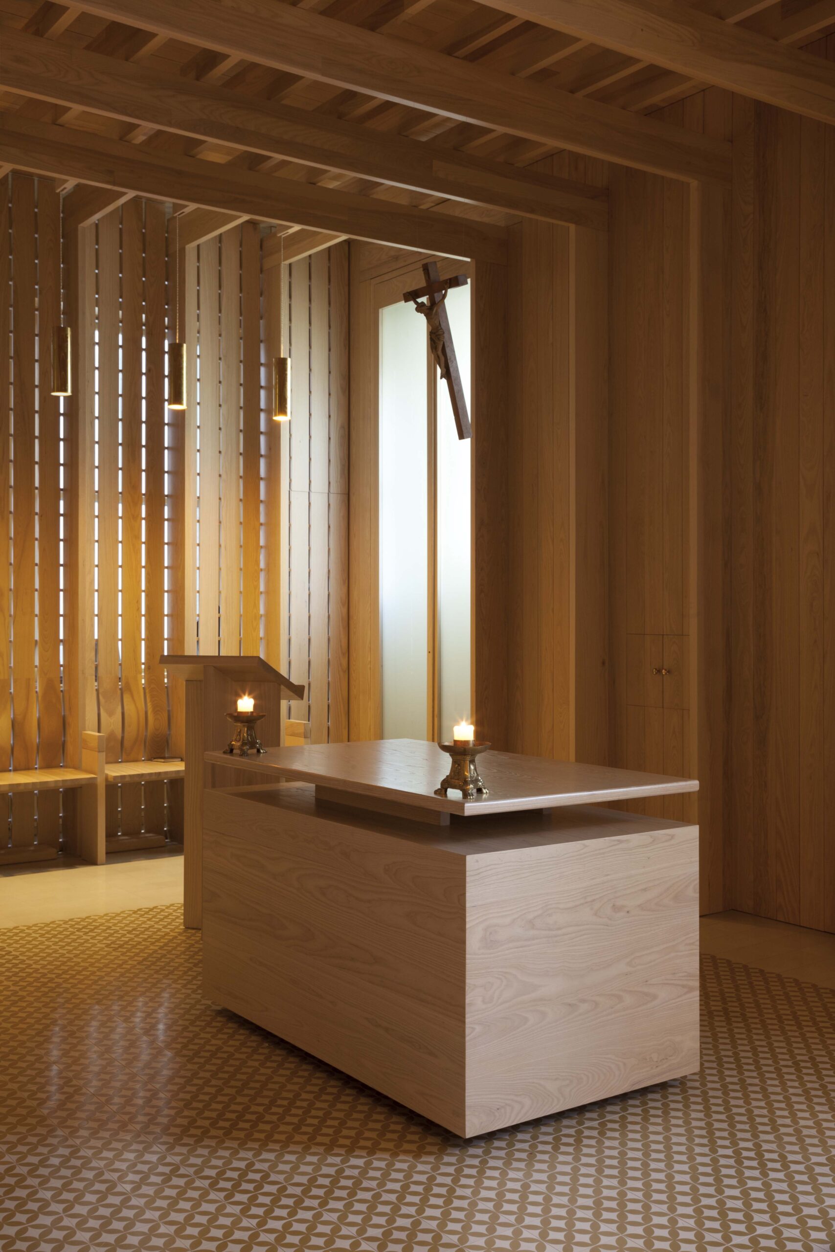 A modern wooden chapel interior, designed in the spirit of Níall McLaughlin, features an altar, two lit candles, a lectern, and a suspended crucifix. Vertical wooden slats and soft lighting create a serene atmosphere.