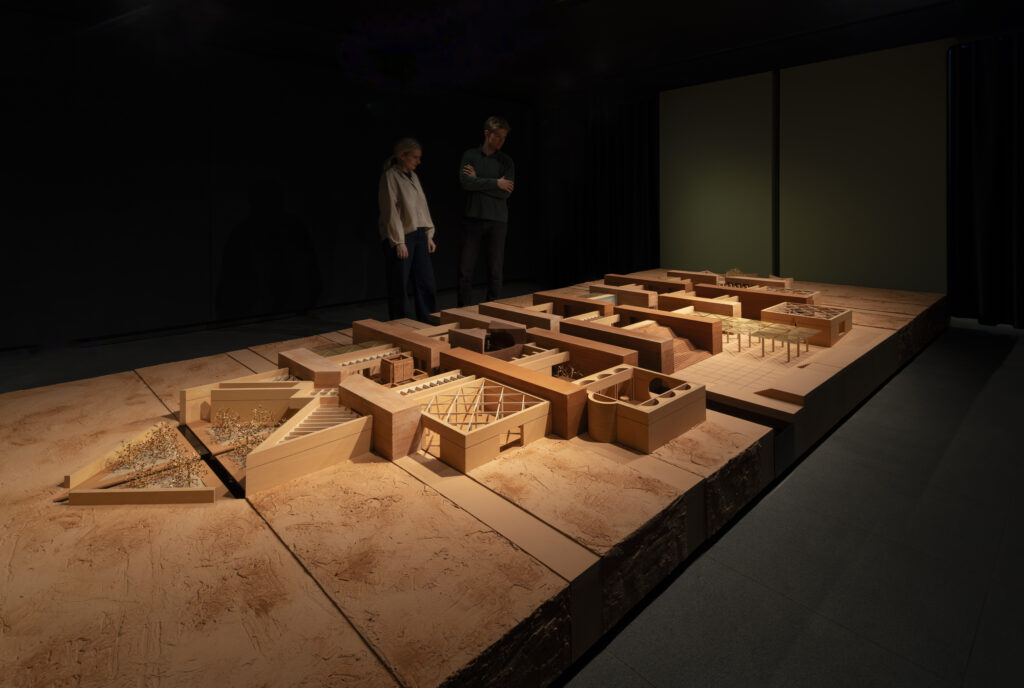 Two people observe a large, detailed architectural model of a Níall McLaughlin building complex displayed on a platform in a dimly lit room.
