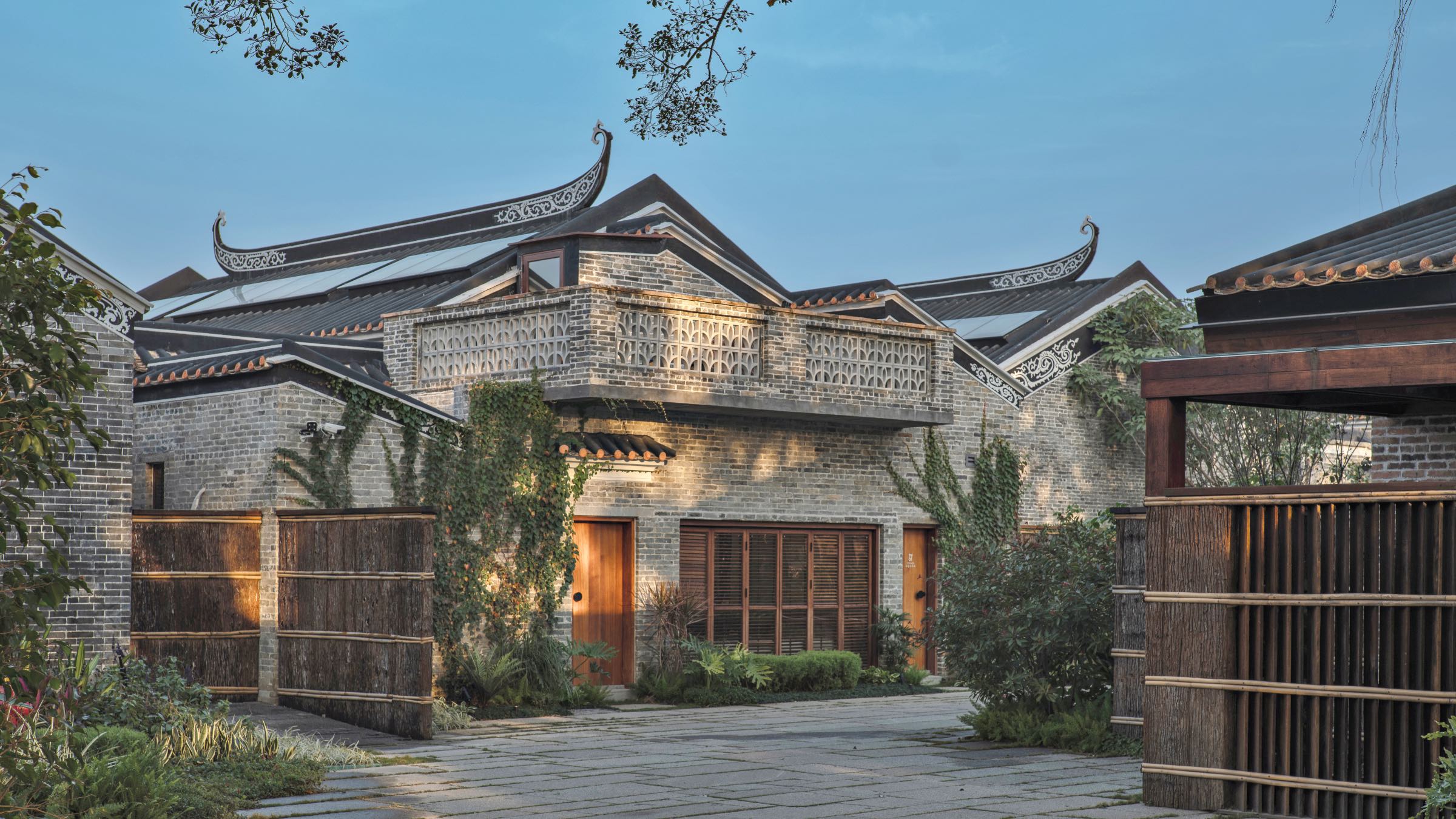 Traditional Chinese brick building by Studio Hu with ornate, upturned roof eaves, wooden doors, and a stone courtyard surrounded by greenery.