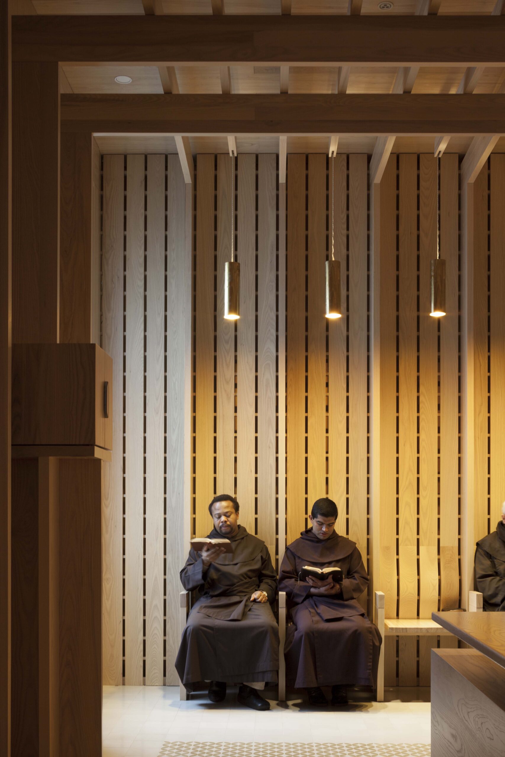 Two men in brown robes sit on a bench in a modern wooden room designed by Níall McLaughlin, each reading a book under hanging pendant lights.