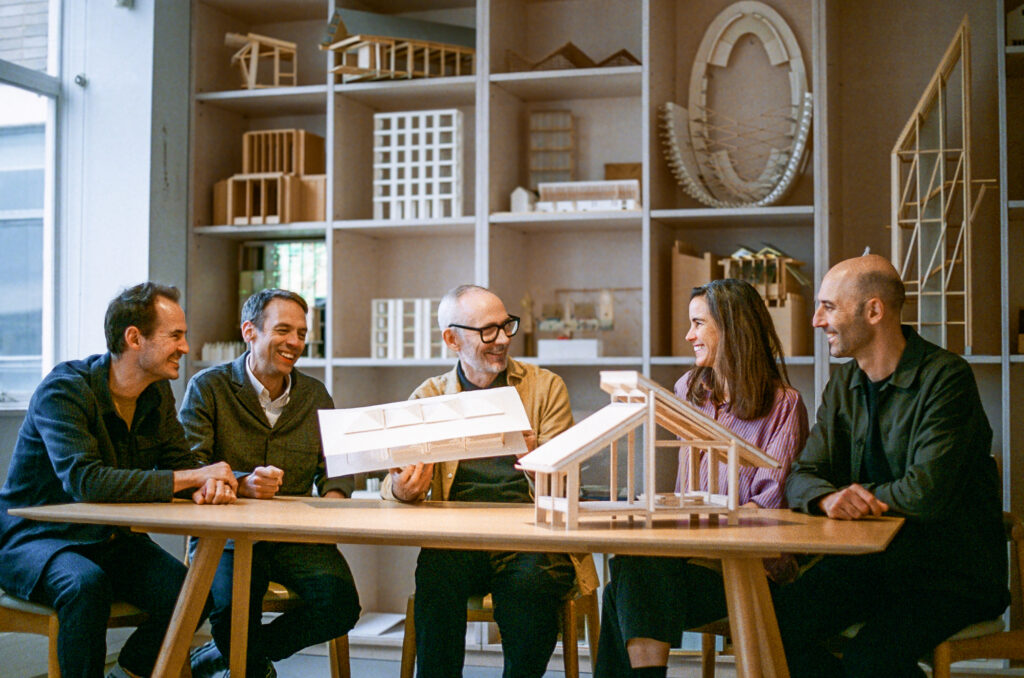 Five people sit at a table, discussing architectural models by Níall McLaughlin, with shelves of wooden structures in the background.