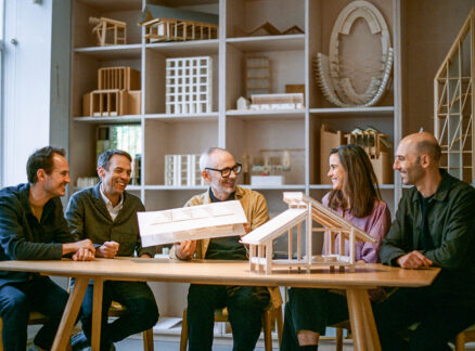 Five people sit at a table, discussing architectural models by Níall McLaughlin, with shelves of wooden structures in the background.