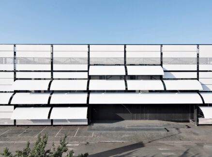 A modern building facade with large white panels, some angled open like shutters, above an empty parking lot under clear blue sky.