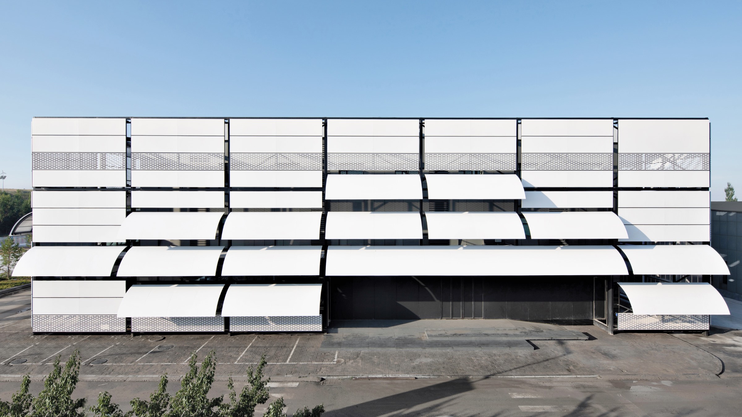 A modern building facade, Erdos Cashmere Centre, with large white panels, some angled open like shutters, above an empty parking lot under clear blue sky.