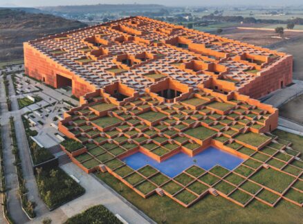 Aerial view of a large, modern building with a geometric, grid-like roof design featuring grass and orange walls, set in a rural landscape.