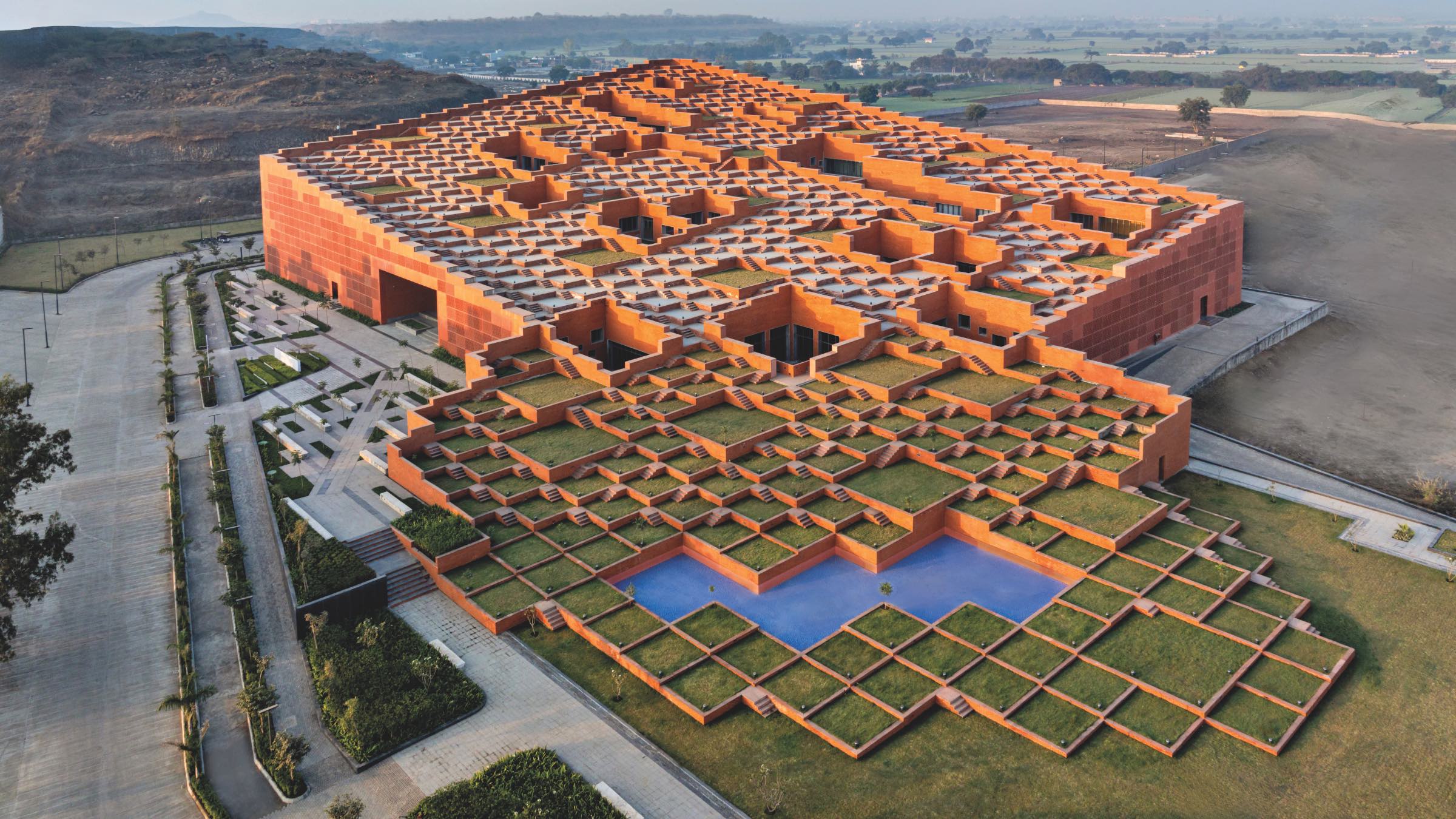 Aerial view of a large, modern building, Prestige University, with a geometric, grid-like roof design featuring grass and orange walls, set in a rural landscape.