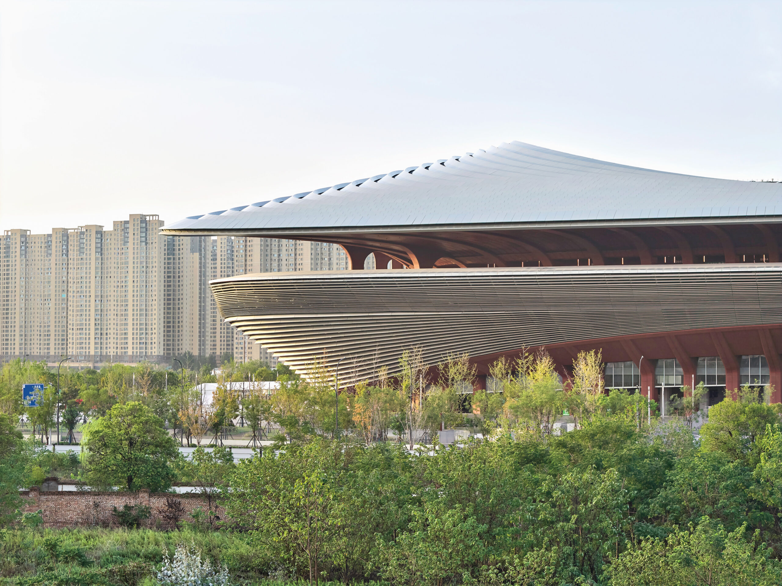 ZHA stadium with a saddle-like form, including a swell in the center and a sweep shelters that spectators by reducing the impact of chilly winds from the north. In the front, greenery and treetops. In the distance, city towers against a light blue sky.