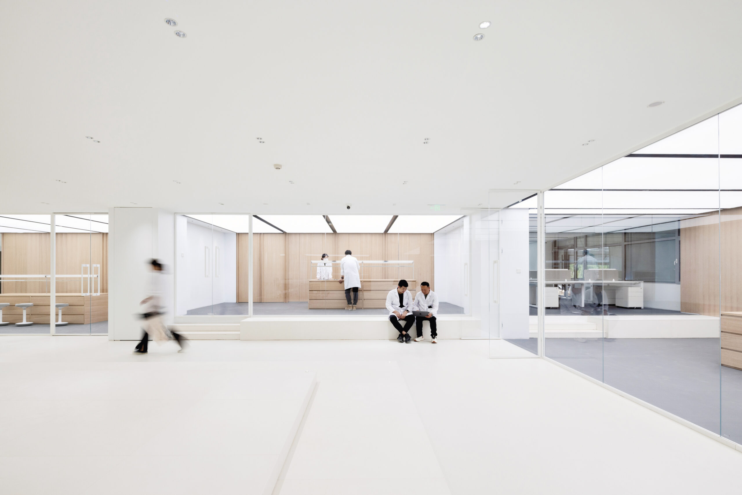 A modern, white, minimalist clinic interior (Erdos Cashmere Centre) with glass walls. Two people in white coats sit on a bench while others walk or stand in the background.
