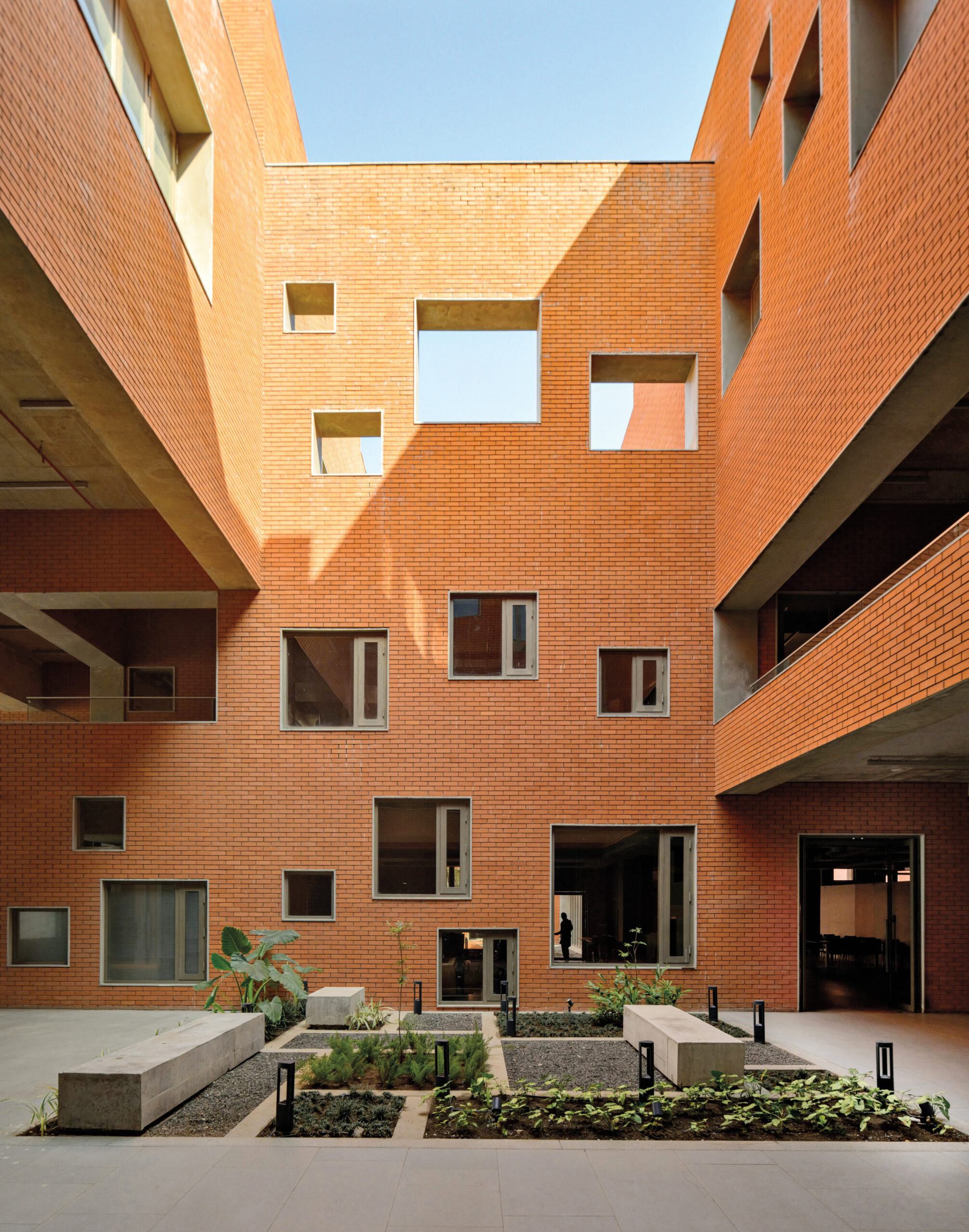 A orange brick wall with multiple windows and cut-outs reveal the blue sky and a multi-floor interior. In front, a gravel and foliage garden with cement seating benches.