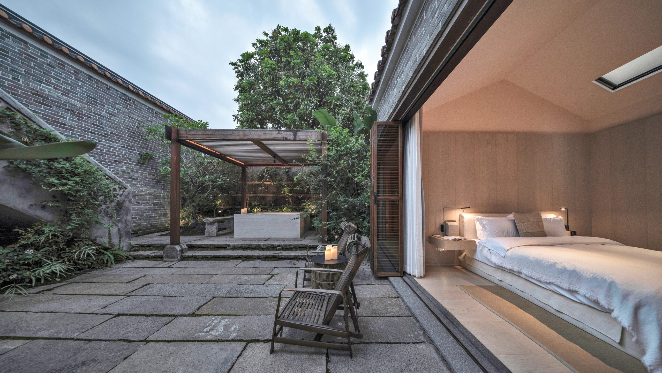 In Langtou Village Inn by Studio Hu, a pale wood bedroom opens onto a stone courtyard with ancient brick walls, greenery, and a seating area.