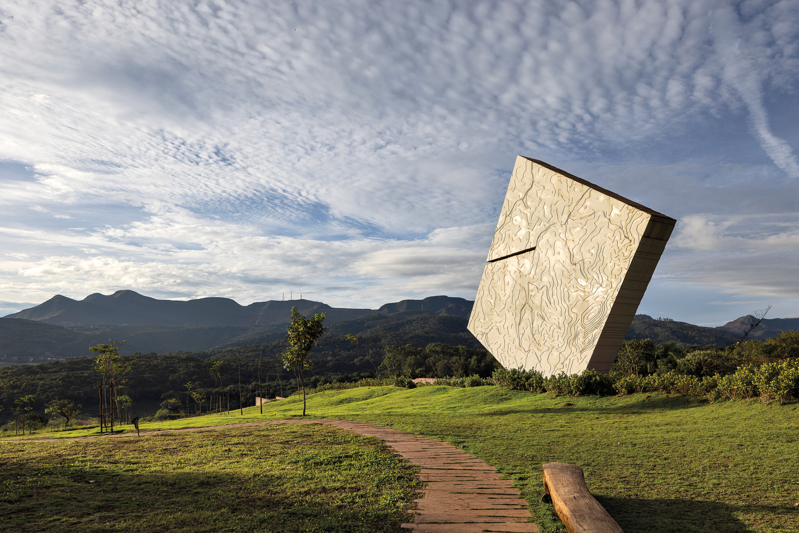 A large, modern, cube-shaped sculpture with a patterned facade stands tilted on a grassy hill, with mountains and a cloudy sky in the background.