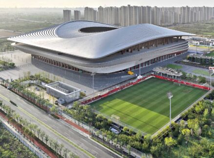 A large modern stadium with a curved roof, surrounded by greenery and smaller football fields, with a city skyline and high-rise buildings in the background.