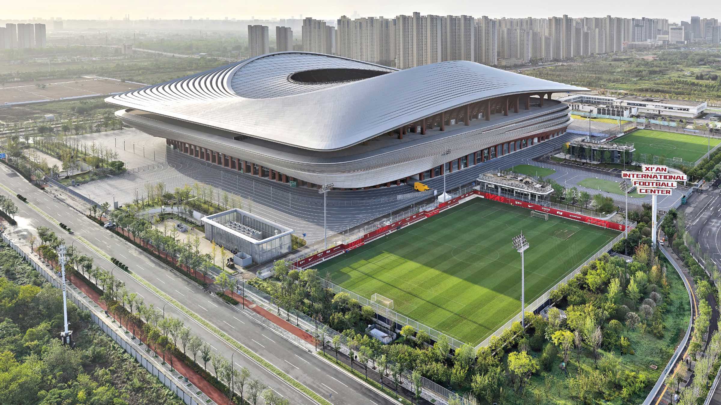 A large modern stadium with a curved roof, surrounded by greenery and smaller football fields, with a city skyline and high-rise buildings in the background.