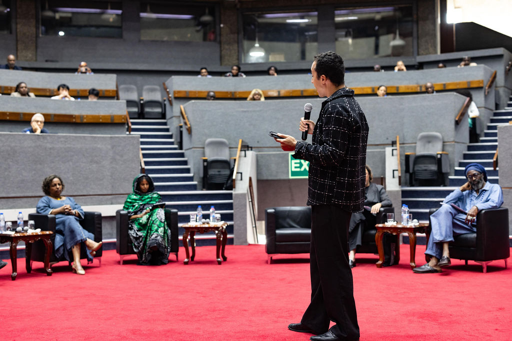 A person stands on a red carpet giving a presentation to a seated audience in an auditorium with gray walls and tiered seating, setting the stage for discussions leading up to UIA 2026 Barcelona.