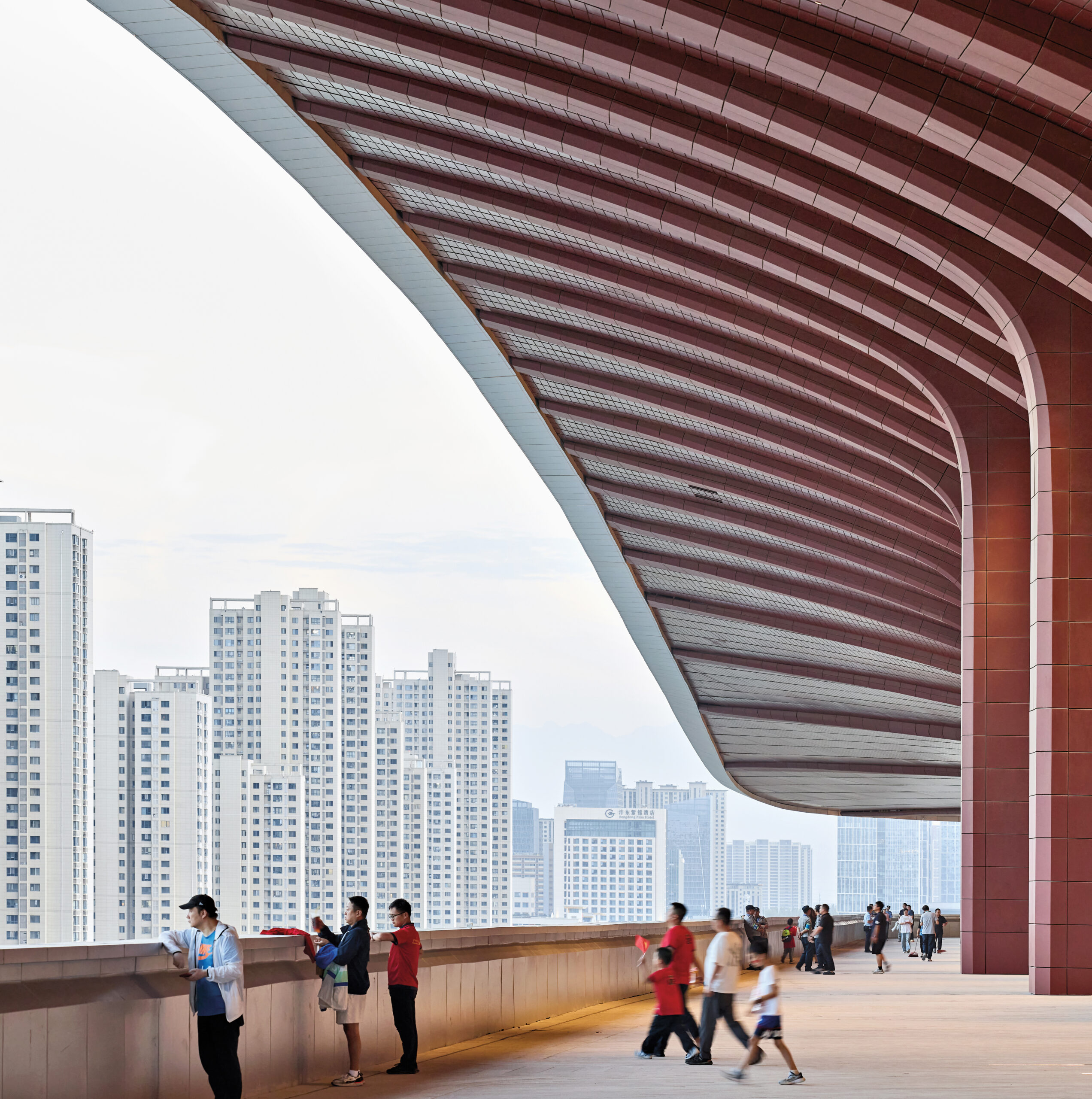 Detail shot of the sweeping roof of the building, with people walking underneath and looking out at the city skyline and urban towers.