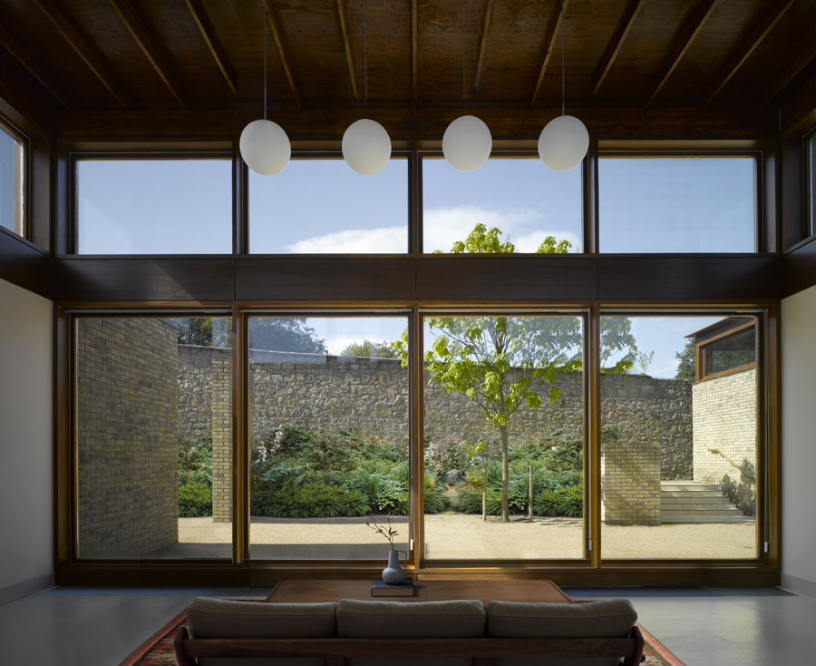 A modern living room with large floor-to-ceiling windows, a sofa, and a view of a stone wall, tree, and outdoor courtyard—evoking the thoughtful design sensibility of Níall McLaughlin. Four round pendant lights hang from a wooden ceiling, adding warmth and elegance to the space.
