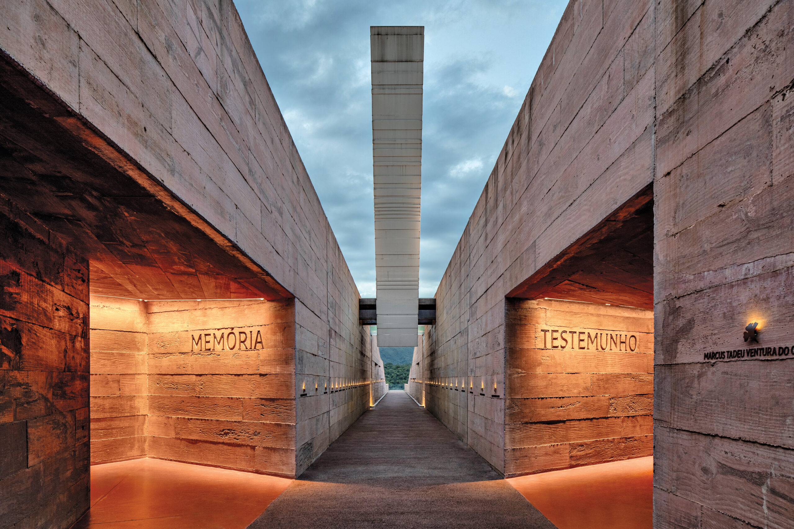 Inside the Brumadinho Memorial: concrete slabs meet textured wood in a linear corridor. On either side, a pathway lit with orange light show the words "Memoria" and "Testemunho".
