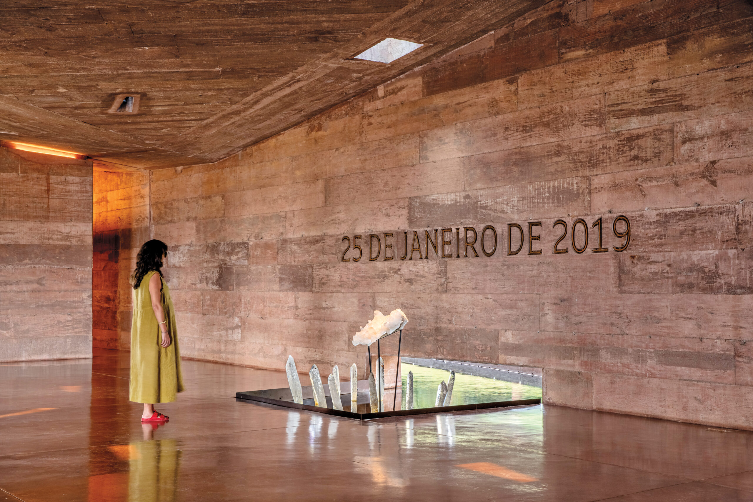 Interior of the Brumadinho Memorial: a woman in a green dress and red sandals stands alone in a raw, stone room looking at a wall that reads, "25 De Janeiro de 2019". Underneath the text, a sculpture of crystal shards sits in a pond that extends outside.