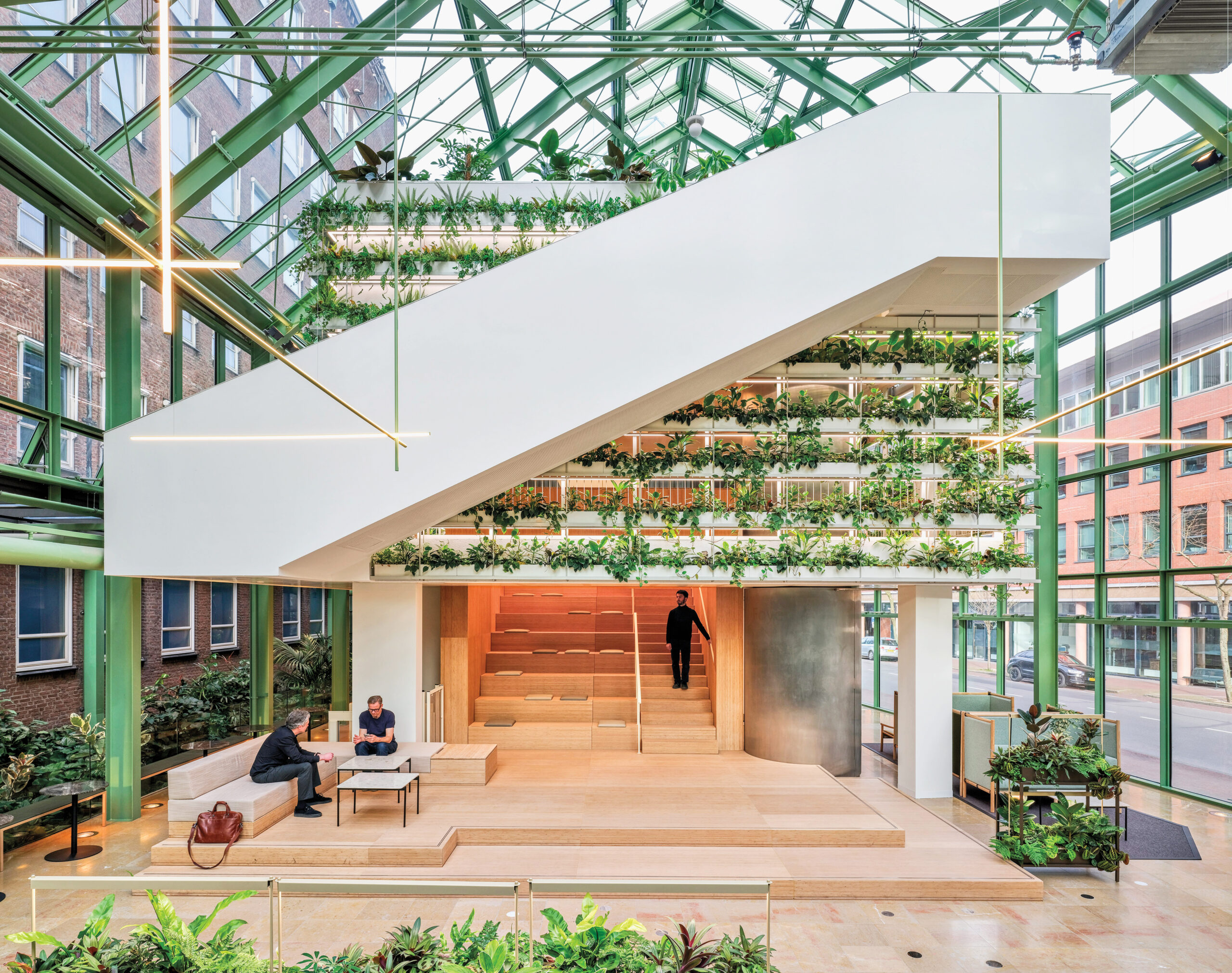 Modern atrium, connecting three Dutch Ministries, with large white staircase, tiered wood seating, abundant plants, glass walls, and two people sitting at a table in the lower left corner.