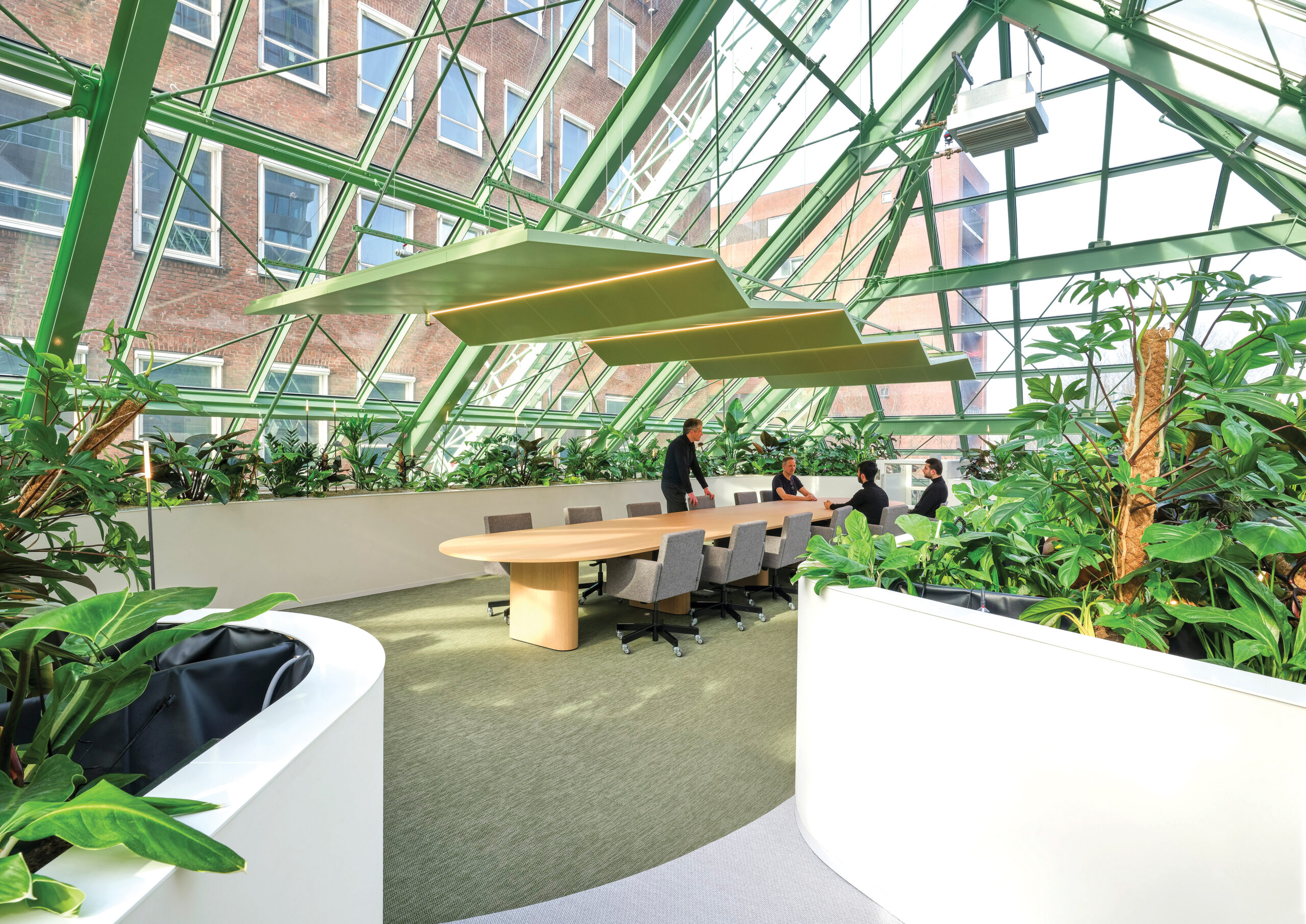 Modern conference room with large wooden table, office chairs, and abundant green plants under a glass roof with geometric framing. Several people stand near the table. Three Dutch Ministries combined.