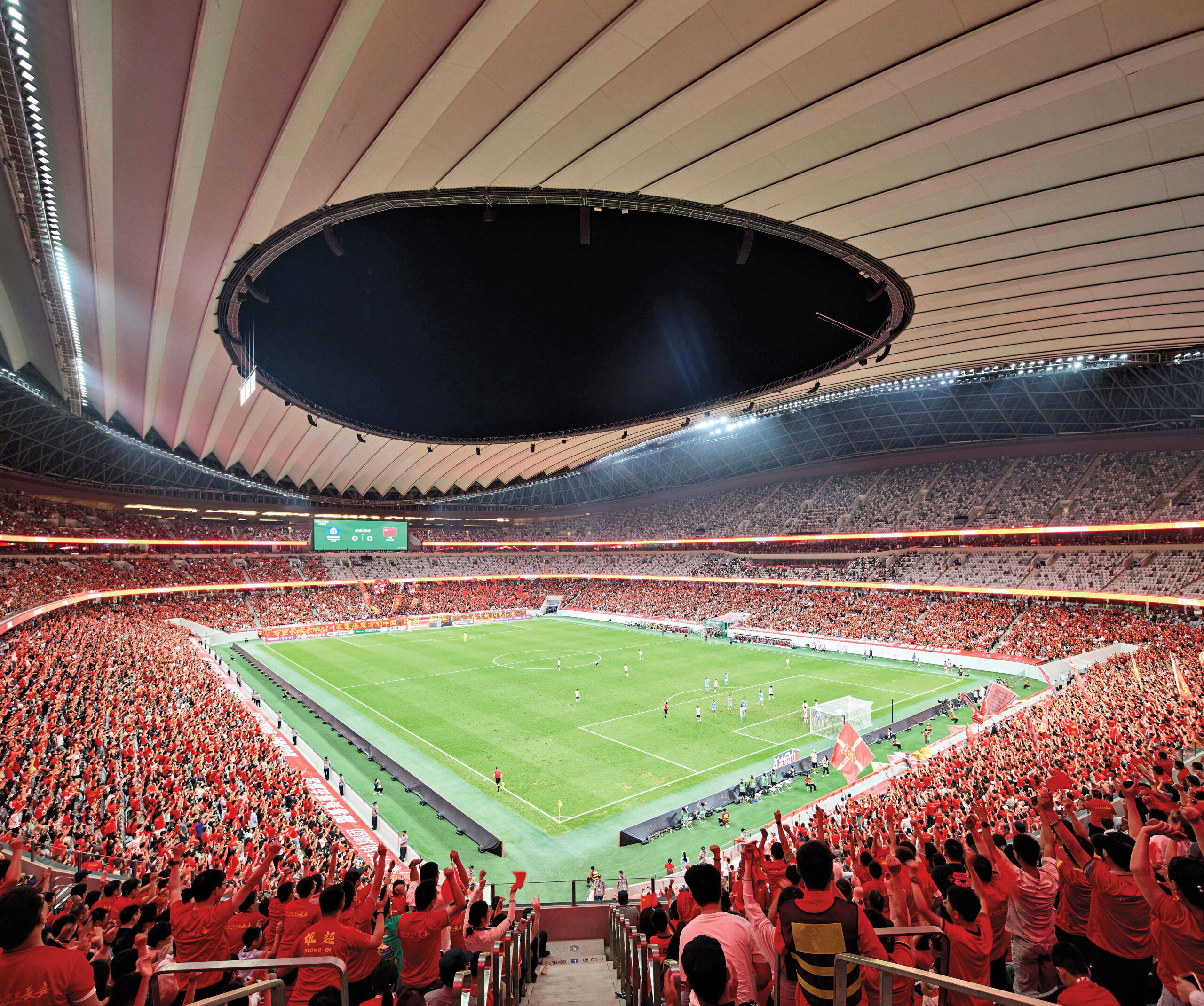 Inside a ZHA stadium with thousands of cheering fans drenched in red light, looking at glowing green football field under a night's sky, with open circular cutout.