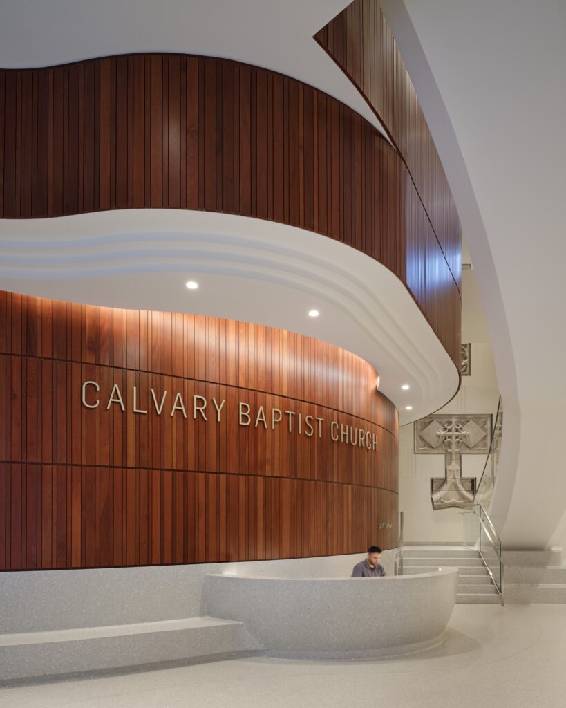 A modern church lobby, inspired by the elegant design sensibility of the Lucas Museum of Narrative Art, features wood paneling, “Calvary Baptist Church” signage, a reception desk, and a staircase with a metal cross displayed on the wall.