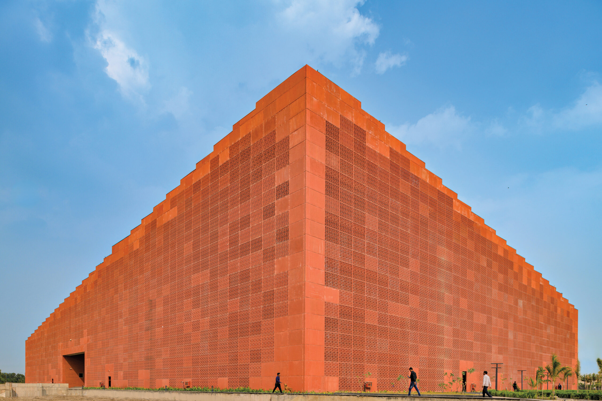 Exterior view of Prestige University: a monolithic orange-clad form against a blue sky, with a few walking figures at the base of the image.