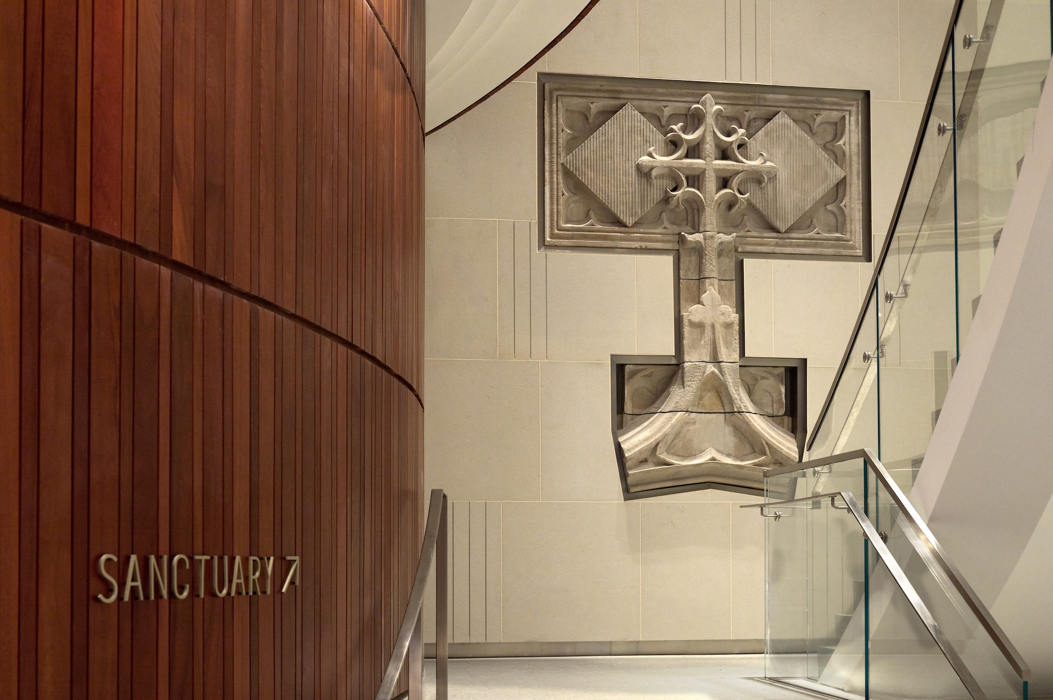 A wooden wall marked “Sanctuary” stands beside a glass stair railing at the Lucas Museum of Narrative Art, near an ornate stone cross embedded in a cream-colored wall.