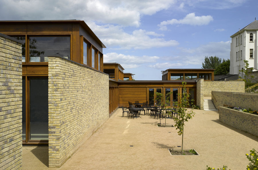 Modern brick and wood building with large windows, outdoor courtyard, tables, chairs, young trees, and a clear sky overhead—echoing the elegant architectural sensibilities of Níall McLaughlin.