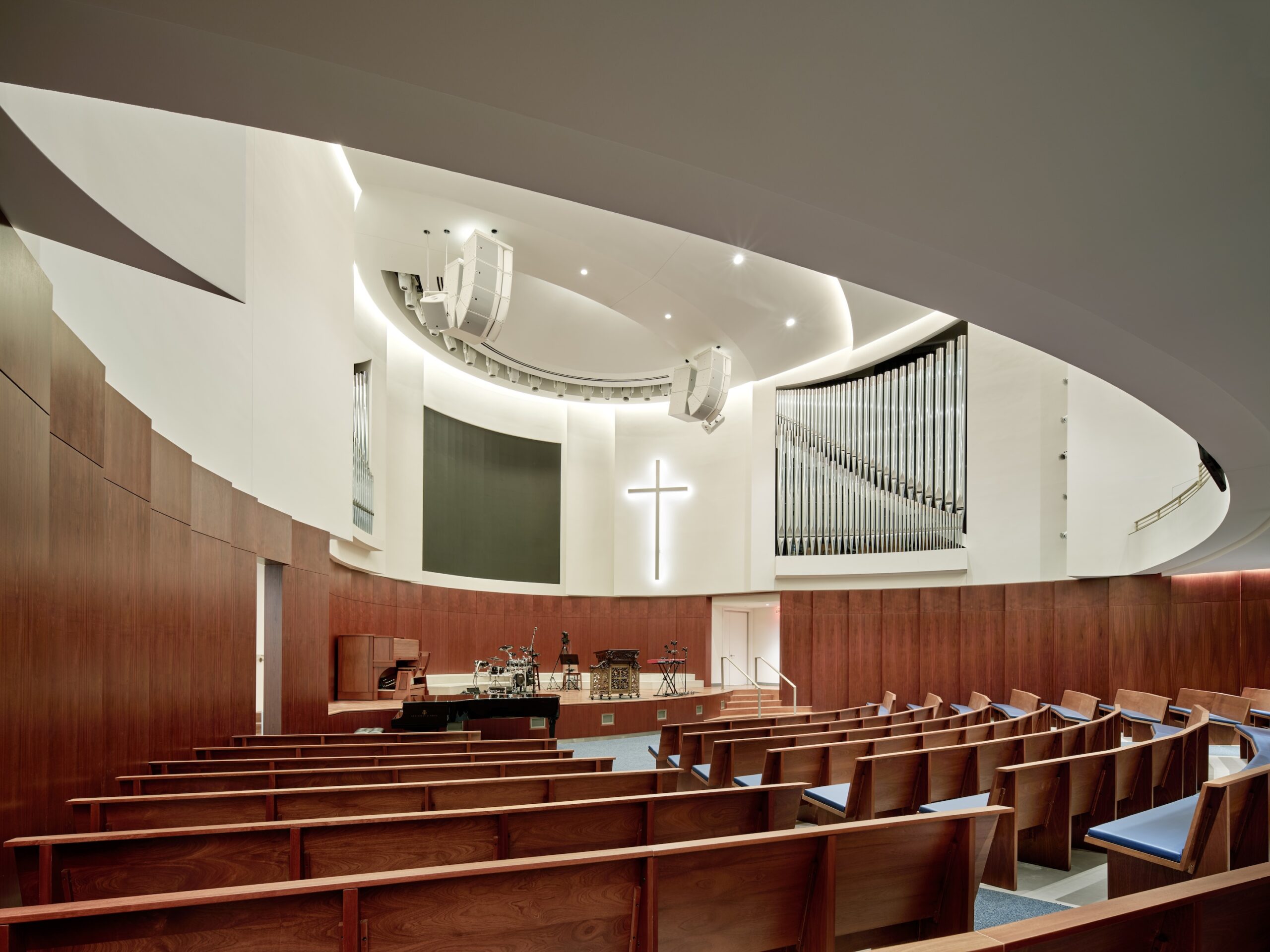 Modern church interior with curved wooden pews, a white illuminated cross on the wall, an organ, and musical instruments on stage under bright lighting—echoing the contemporary design sensibilities of the Lucas Museum of Narrative Art.