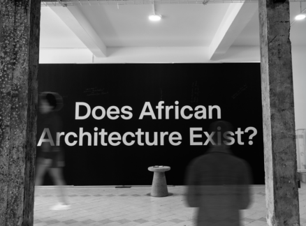 A black wall with large white text reading “Does African Architecture Exist?” is displayed behind two blurred figures and a small stool, evoking the thought-provoking spirit anticipated at UIA 2026 Barcelona.