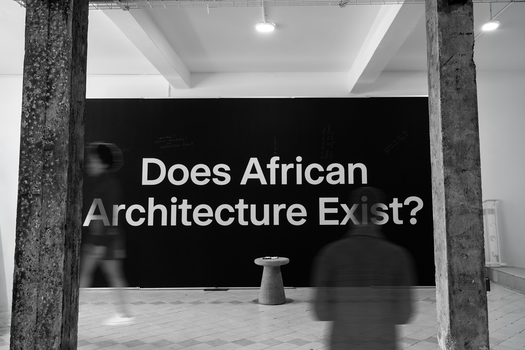 A black wall with large white text reading “Does African Architecture Exist?” is displayed behind two blurred figures and a small stool, evoking the thought-provoking spirit anticipated at UIA 2026 Barcelona.
