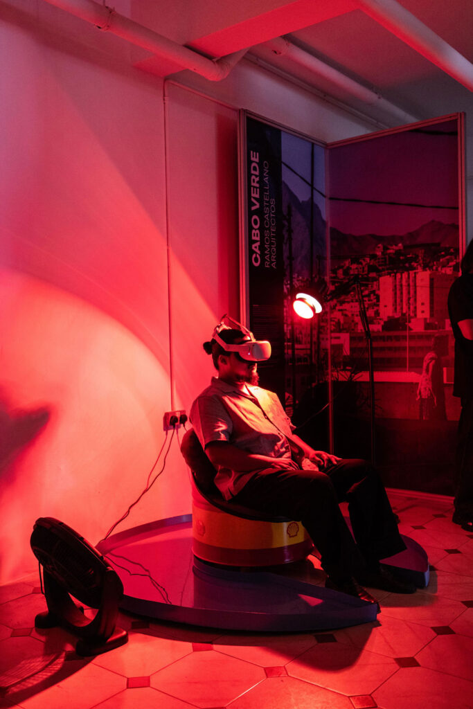 A person sits in a chair wearing a VR headset under red lighting in a room with tiled flooring, with a Cabo Verde promotional panel and UIA 2026 Barcelona signage visible in the background.