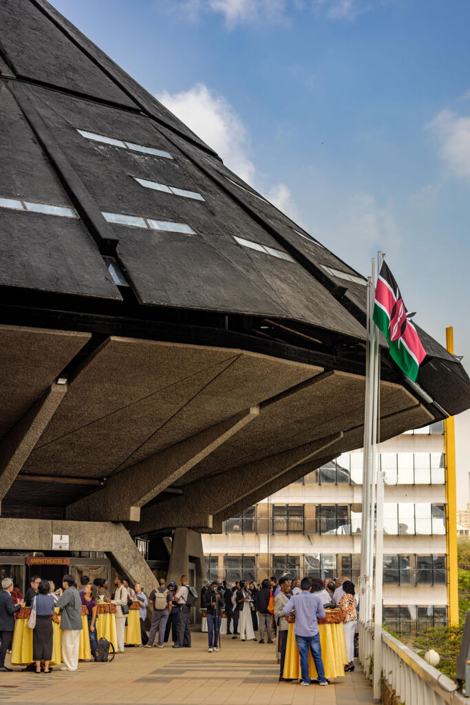 People gather outside a modern building with a sloped concrete roof, next to a Kenyan flag, under a partly cloudy sky—an atmosphere reminiscent of the vibrant architectural exchanges anticipated at UIA 2026 Barcelona.