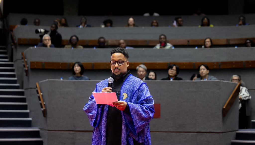 A person in a blue robe stands and reads from a pink paper, addressing an audience seated in tiered rows inside an auditorium during the UIA 2026 Barcelona event.