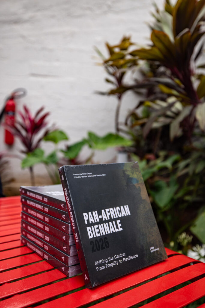 A stack of books titled “PAN-AFRICAN BIENNALE 2026” sits on a red table with green plants in the background, subtly reflecting the global spirit of events like UIA 2026 Barcelona.