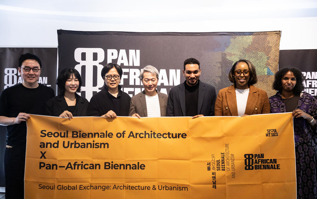 Eight people stand in a row holding a yellow banner for the Seoul Biennale of Architecture and Urbanism X Pan-African Biennale, in front of a backdrop with event logos, highlighting their connection to UIA 2026 Barcelona.