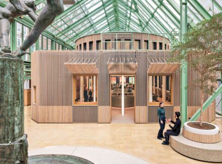 Building Common Ground Across Three Dutch Ministries Interior of a modern glass atrium with a central wooden structure, two people conversing near a planter, and a bronze sculpture in the foreground.