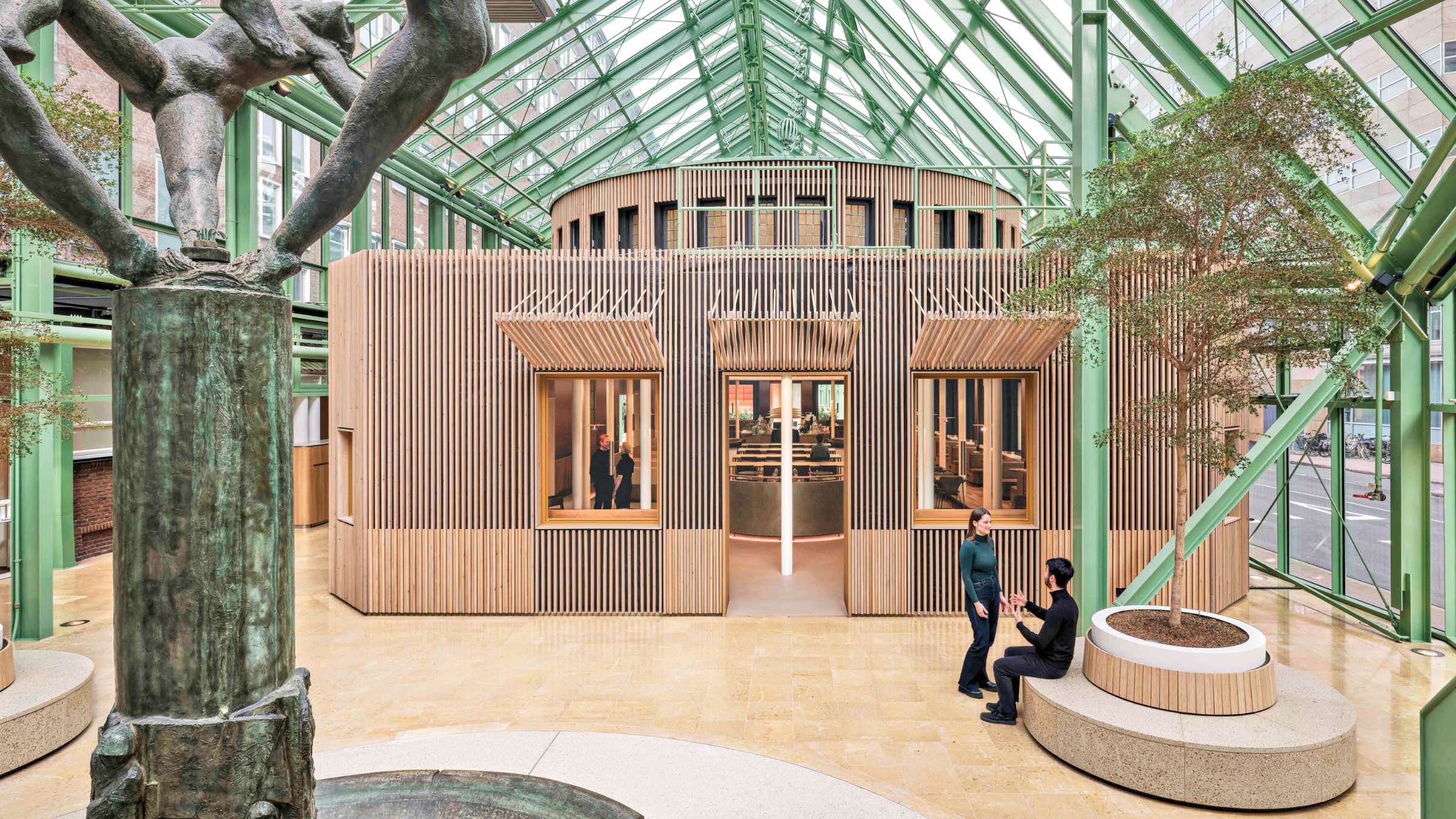Interior of a modern glass atrium with a central wooden structure, two people conversing near a planter, and a bronze sculpture in the foreground. Connecting 3 Dutch Ministries.