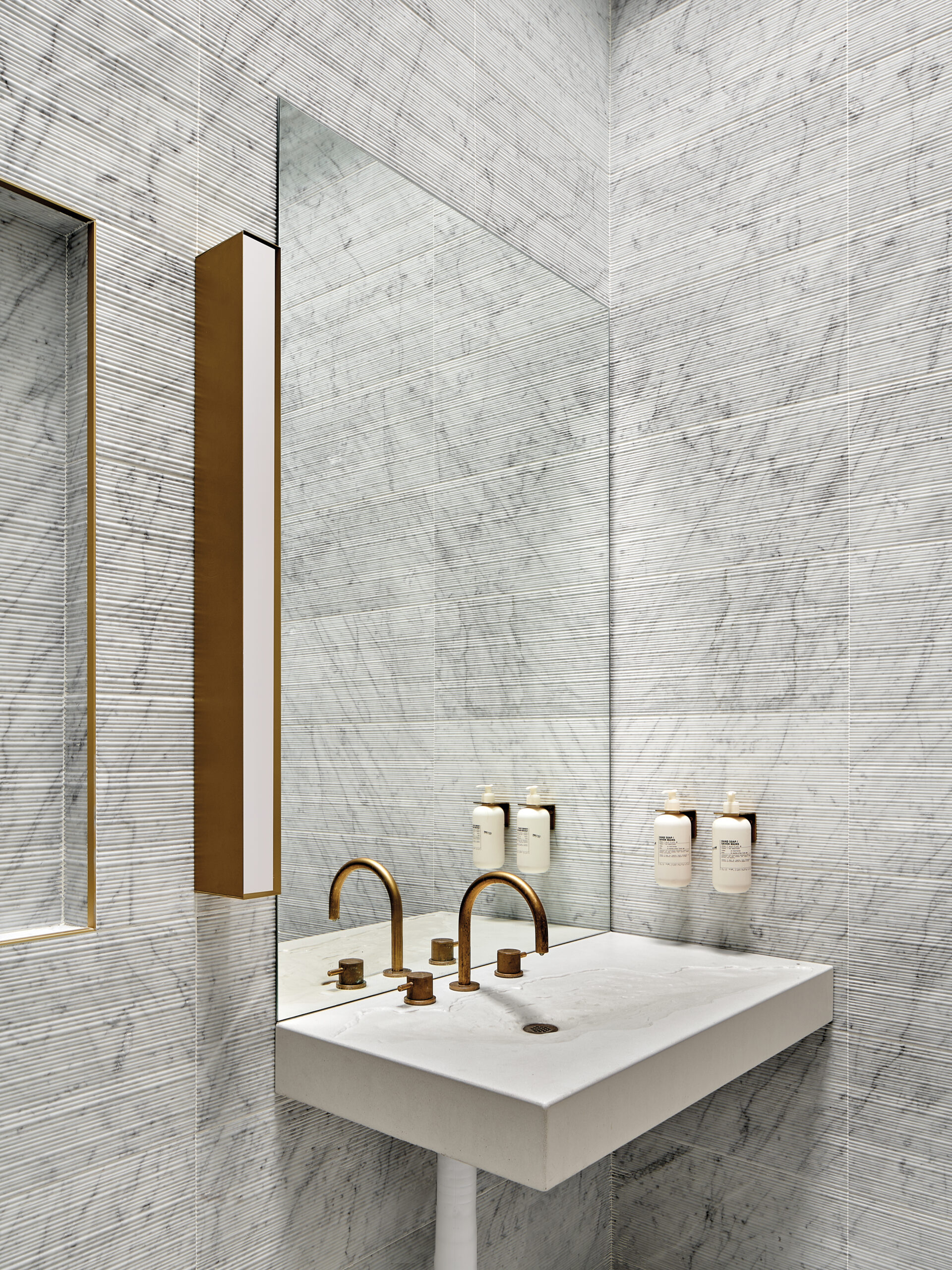 Products to Specify: A modern white sink in a grey bathroom with a textural wall surface and brass finishes.