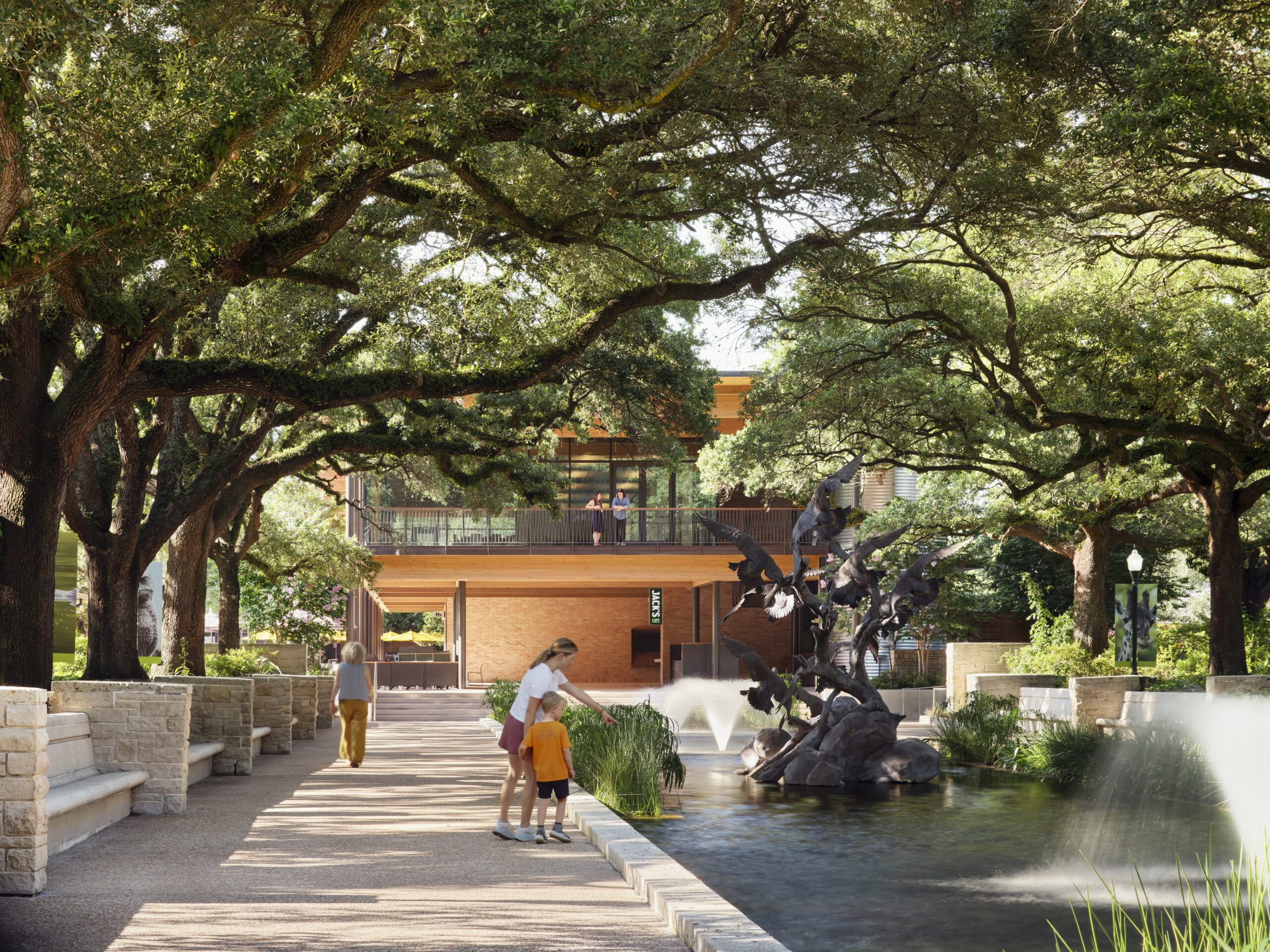 A woman and child stand by a fountain with a sculpture, surrounded by large trees and the Lucas Museum of Narrative Art in the background.