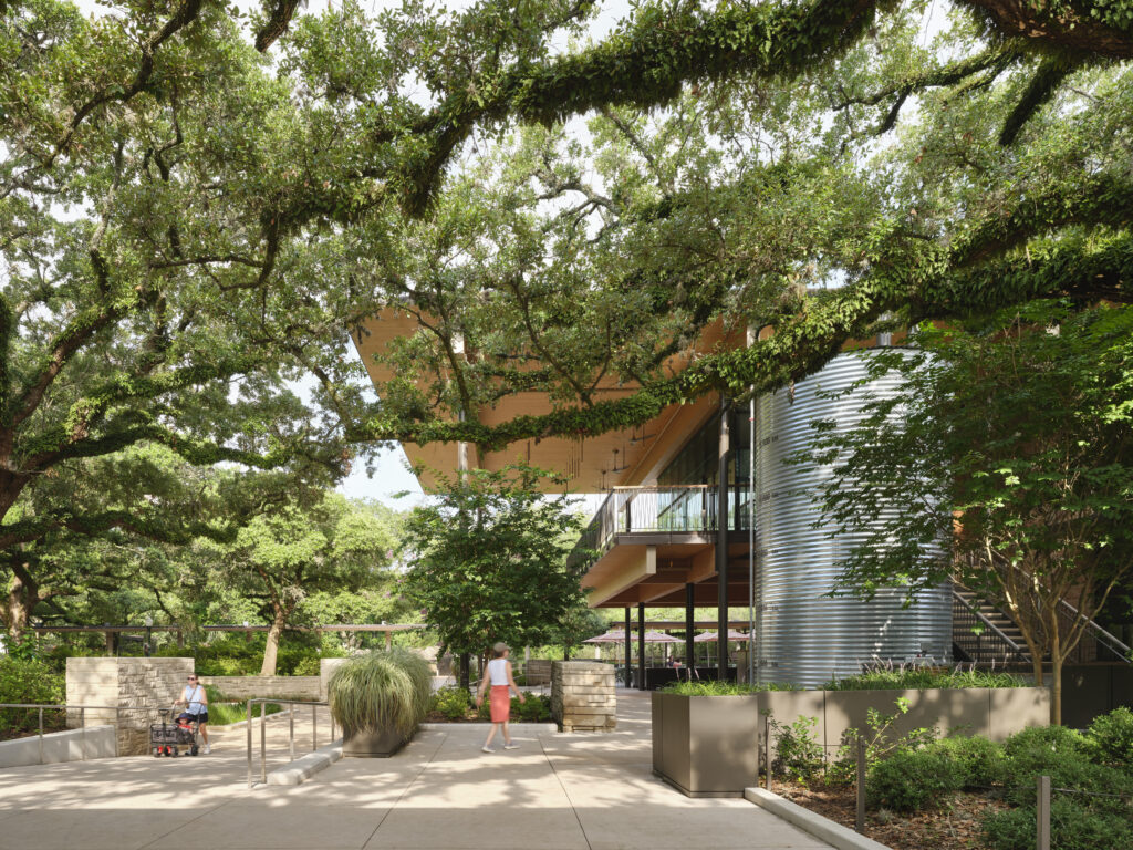 A person walks along a shaded path near the modern Lucas Museum of Narrative Art, with its large windows and metal siding, surrounded by trees and lush greenery.