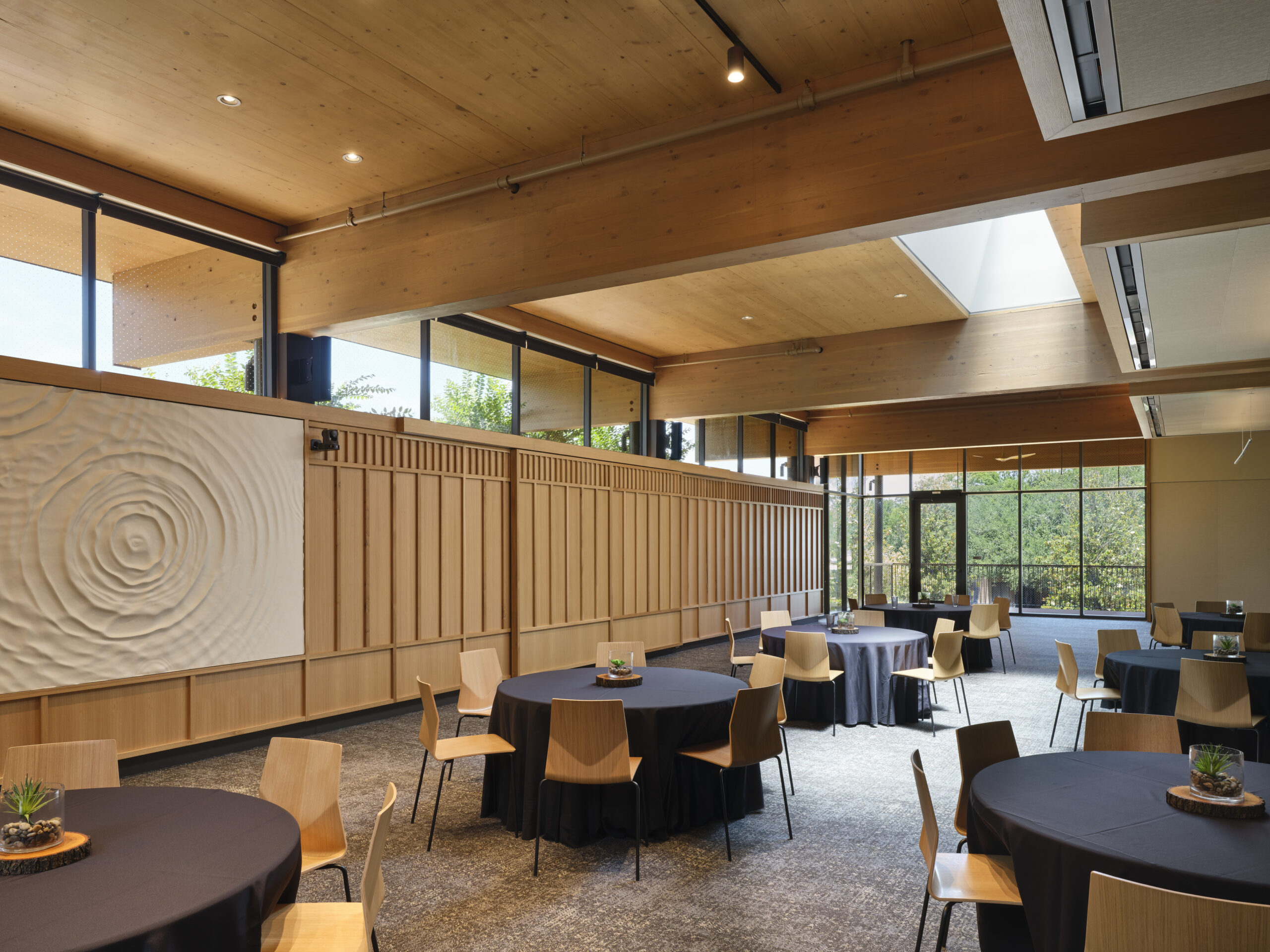 Spacious event room at the Lucas Museum of Narrative Art with round tables covered in black cloth, wooden chairs, large windows, wood ceiling, and a textured white wall art panel.