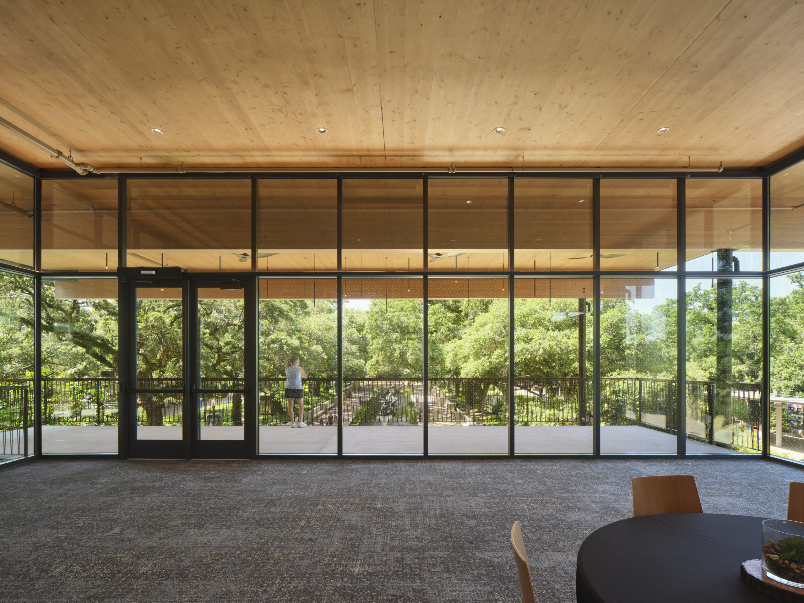 A large glass wall reveals a view of trees and a wooden deck outside the Lucas Museum of Narrative Art; a person stands on the deck, and empty chairs and tables are visible inside the room.