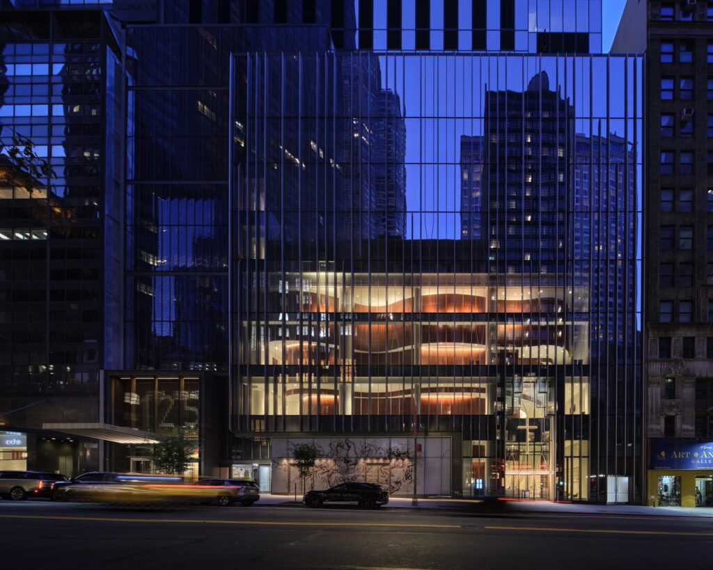 Modern glass building at dusk with illuminated interior and reflections of surrounding skyscrapers; reminiscent of the sleek design of the Lucas Museum of Narrative Art, with cars and light trails visible on the street in front.