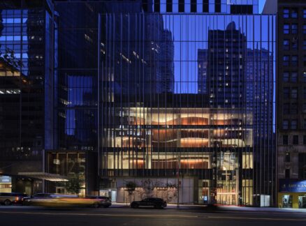 Modern glass building at dusk with illuminated interior and reflections of surrounding skyscrapers; reminiscent of the sleek design of the Lucas Museum of Narrative Art, with cars and light trails visible on the street in front.